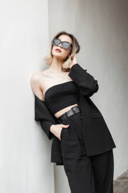 Stylish beautiful elegant girl in fashionable black clothes with trendy top, fashion black blazer and pants standing near the white wall on the street. Women's Urban Business Style