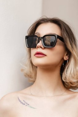 Beautiful female headshot portrait of a pretty stylish young girl with a nose piercing with black fashion sunglasses on a white background on the street