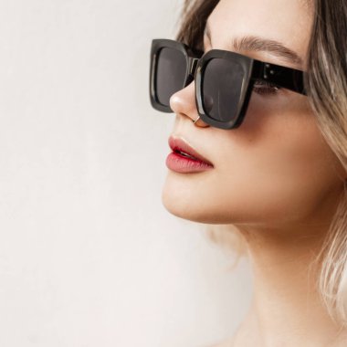 Woman's close-up portrait of a beautiful young girl with nose piercing and lips with black fashionable modern sunglasses on white background outdoors. Adorable female headshot.