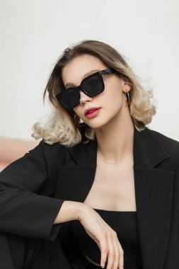 Vogue elegant business fashion beauty woman with fashionable sunglasses in black outfit with top and blazer sits on the street