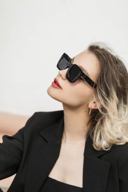 Street female portrait of beautiful young woman model with trendy fashion sunglasses in black clothes with blazer and top sits and rest on the street