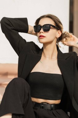 Fashionable beautiful young woman with cool fashionable sunglasses in stylish black elegant clothes sits and poses on the street