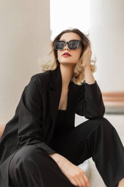 Cool elegance fashionable young pretty lady model with sunglasses in black business fashion clothes with stylish blazer sits on the street near a white column