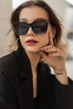 Fresh urban female portrait of business young stylish trendy woman model with cute face with stylish modern sunglasses in black elegant clothes with blazer sits and poses in the city