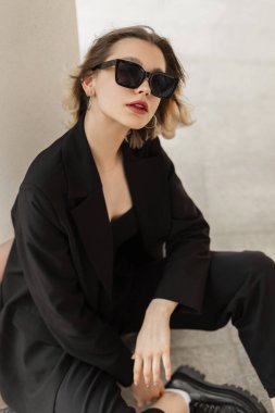 Fashionable young woman model with black elegance business clothes with blazer and sunglasses sits on the street near a white wall