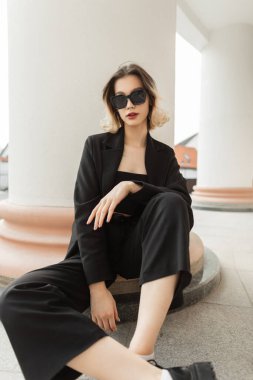 Fashionable young beautiful business woman model with trendy modern sunglasses in black elegant clothes with blazer, top and pants sits near a vintage column in the city