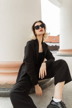 Fashionable young business stylish beautiful woman model with black fashion sunglasses in black trendy elegance outfit with blazer, top and pants sits and poses near a white vintage column in city