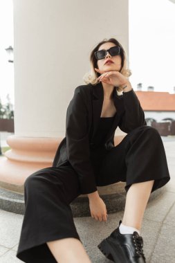 Fashionable elegance vogue pretty woman with sunglasses in black business fashion clothes with black blazer and pants with shoes sits on the street near a vintage white column