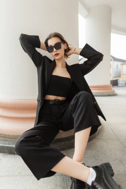 Fashionable young pretty business woman model with trendy modern sunglasses in black elegant clothes with blazer, top and pants sits near a vintage column in the city