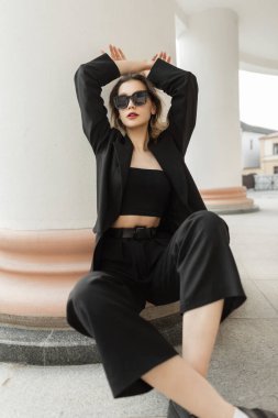 Fashionable elegance vogue pretty woman with sunglasses in black business fashion clothes with black blazer and pants with shoes sits on the street near a vintage white column