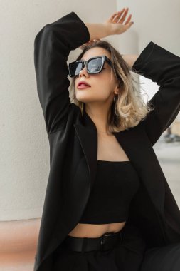 Fashion adorable beautiful young elegance woman model with stylish sunglasses in black business fashionable clothes with blazer and top sits and poses in the city near a vintage white column