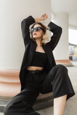Fashion adorable beautiful young elegance woman model with stylish sunglasses in black business fashionable clothes with blazer and top sits and poses in the city near a vintage white column