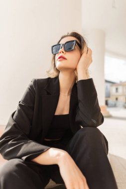 Trendy business fashion beauty woman model with black stylish sunglasses in black urban elegant outfit with top, blazer and pants sits in the city near a white vintage column