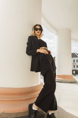 Elegant vogue beautiful young business woman model with cool trendy sunglasses in fashion strict black outfit with jacket, pants, top and shoes standing near a white vintage column in the city