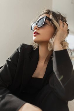Fashion glamour pretty lade woman with modern sunglasses in black elegant business clothes sits and rest on the street