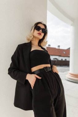 Fashionable elegant trendy beautiful woman model with modern sunglasses in black business look outfit with fashion blazer, top and pants stands near a white column in the city