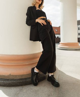 Beautiful stylish young woman model in black fashionable business outfit with a jacket, top, pants and leather shoes stands near a white vintage column in city. Women's elegant urban style and fashion