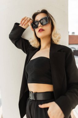 Fashionable pretty elegant business girl in fashionable black clothes wears sunglasses posing near a white vintage column