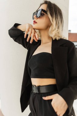 Fashionable elegant trendy beautiful woman model with modern sunglasses in black business look outfit with fashion blazer, top and pants stands near a white column in the city