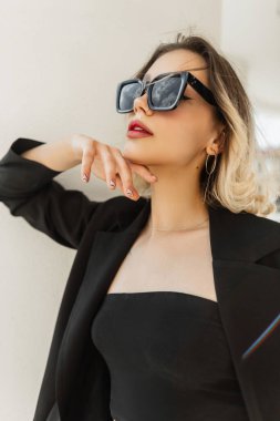 Beautiful young elegant stylish woman model hipster in fashionable black sunglasses with black business blazer and top standing near the white wall on the street