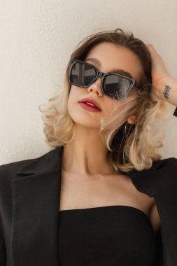 Beautiful young elegant stylish woman model hipster in fashionable black sunglasses with black business blazer and top standing near the white wall on the street
