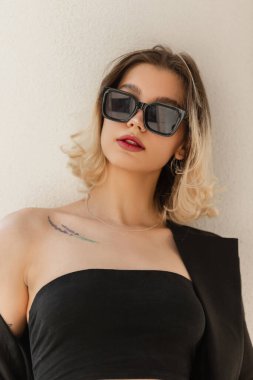 Woman's fresh urban portrait of a beautiful young woman with short hair with cool black sunglasses and a black top with blazer stands near a white wall in the city
