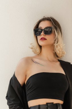 Stylish beautiful young fashionable business successful woman with cool glasses in fashion strict elegant clothes with blazer and top near white wall in the city. Women's fashion, beauty and style