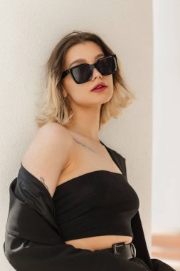 Urban female pretty stylish vogue portrait of beautiful young model woman with short hair with trendy modern sunglasses in black business elegant clothes look with blazer and top stands near a white wall