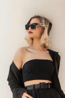 Urban female stylish vogue portrait of beautiful young model woman with short hair with trendy modern sunglasses in black business elegant clothes look with blazer and top stands near a white wall