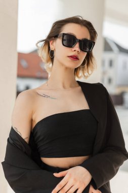 Fashionable young trendy elegance woman model with fashion modern sunglasses in stylish black business clothes with top and blazer walks in the city in sunny summer day