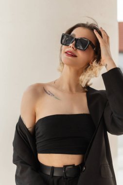 Fashionable happy young vogue beauty lady with smile with fashion sunglasses in trendy black business look outfit with top, blazer and pants walks in the city in sunny day