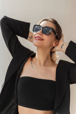 Happy beautiful elegant young model girl with a smile and cool sunglasses in fashion black business clothes with a trendy top and blazer walking in the city on a sunny day. Natural feminine beauty