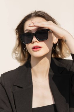 Fashionable beautiful business lady model with modern sunglasses in fashion black elegant clothes with stylish blazer sits in the sunny day and covers her eyes with hand. Summer female urban look