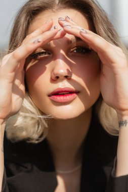 Women's summer fresh portrait of a beautiful young elegant woman model with a short haircut sits in the street, covering eyes with her hands from the sunlight and looking into the camera in sunny day