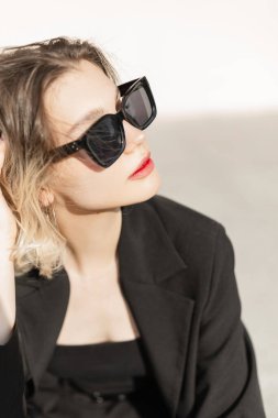 Summer female fresh portrait of beautiful young woman model with cool trendy sunglasses in black elegant clothes sits outdoors at sunlight