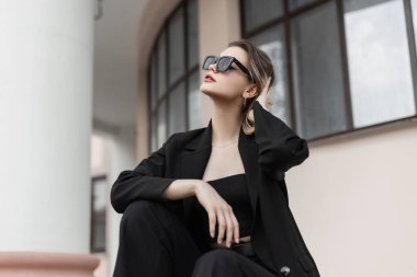 Stylish beautiful young woman model with black sunglasses in fashion black business outwear with a jacket sits in the city. Female trendy elegant clothes look