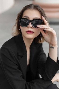 Elegance fashion stylish business beauty woman in black fashionable clothes with blazer wears a trendy modern sunglasses and sits on the street