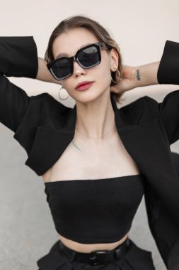 Stylish urban beautiful young woman model with fashionable sunglasses in fashion black elegant outfit with a blazer and a top sitting in the city. Street fashion vogue female clothes look