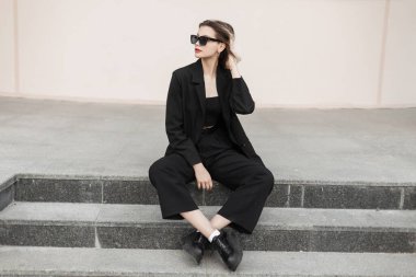 Stylish beautiful elegant model girl with fashion cool sunglasses in fashionable black business clothes with top, blazer, pants and shoes sits on the steps of the street. Vogue woman
