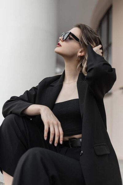 Fashionable beautiful young business lady with fashionable sunglasses in a fashion black blazer with a top and pants is sitting outside and fixing her hair. Elegant women's style and beauty