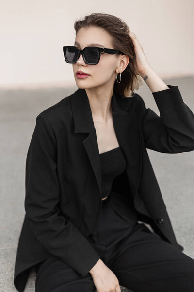 Модная городская женщина свежей красоты в модных солнцезащитных очках в черном бизнесе элегантная одежда с блейзером и топ с брюками сидит на улице