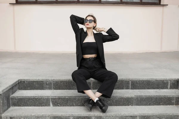 Fashion business pretty girl with sunglasses in a fashionable black elegant suit outfit with jacket, top, pants and shoes sits on the steps outside the building