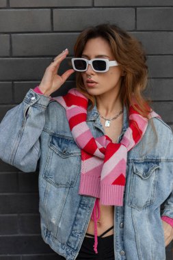 Şık, genç, güzel, kızıl saçlı hippi kadın. Şık şehir kıyafetleri içinde, kot pantolon, pembe bir sweatshirt ve üstü modern güneş gözlüğü takıyor ve şehirde siyah tuğlalı bir duvarın yanında duruyor.