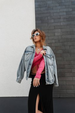 Güzel, modaya uygun, beyaz güneş gözlüklü, şık şehir kıyafetleri içinde, kot ceketli, pembe bluzlu ve uzun etekli bir kadın modeli sokakta bir duvarın yanında duruyor.