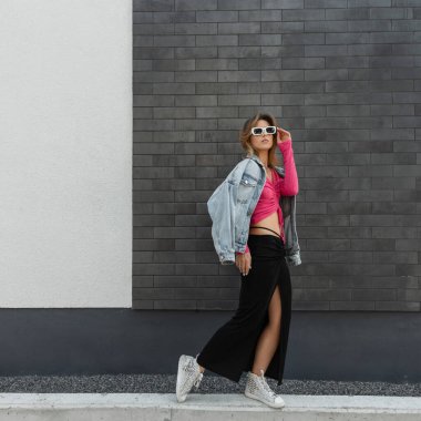 Havalı moda güzel hippi kız şık parlak giysiler içinde kot ceket, pembe bluz, elbise ve ayakkabılar giyip şık beyaz güneş gözlüğü takıyor ve sokakta duvarın yanında yürüyor.