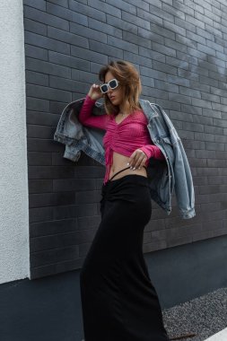 Pembe bluzlu, uzun etekli ve kot ceketli şık, genç ve güzel bir bayan model hippi şık bir beyaz gözlük takıyor ve siyah bir duvarın yanında poz veriyor