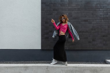 Havalı, beyaz güneş gözlüklü, şık, moda ceketli, pembe bluzlu, etekli ve sokakta duvarda dans eden spor ayakkabılı şık, modaya uygun bir hippi kadın.