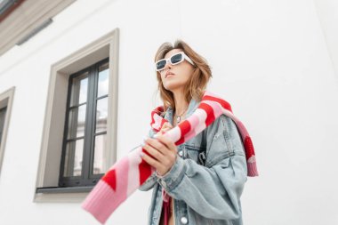 Beyaz güneş gözlüklü, şık, kot ceketli, pembe örülü sweatshirt 'lü güzel, kızıl saçlı hippi kadın sokakta beyaz bir binanın yanında yürüyor.