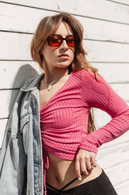 Güzel, seksi bir kadın model. Güneş gözlüklü, havalı bir kot ceketli, pembe etekli. Güneşli bir günde plajda, eski moda beyaz bir duvarın yanında duruyor.