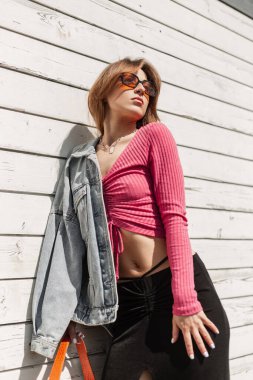Güzel, turuncu gözlüklü, havalı, günlük kot elbiseli, pembe ceketli, etekli ve çantalı güzel moda kadını güneşli bir günde klasik beyaz ahşap duvarın yanında duruyor.